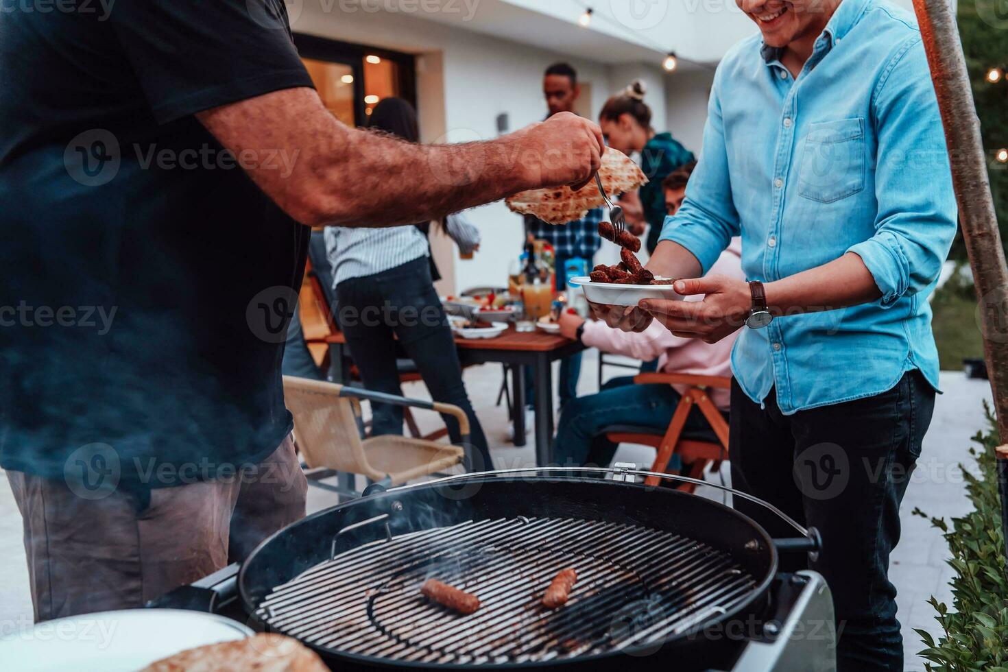 A group of friends and family barbecue together in the evening on the terrace in front of a large modern house photo