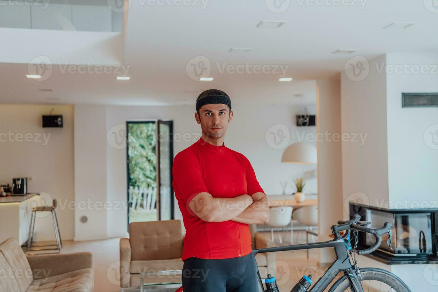 Photo of a determined triathlete standing in a modern large living room, behind him is his training bike