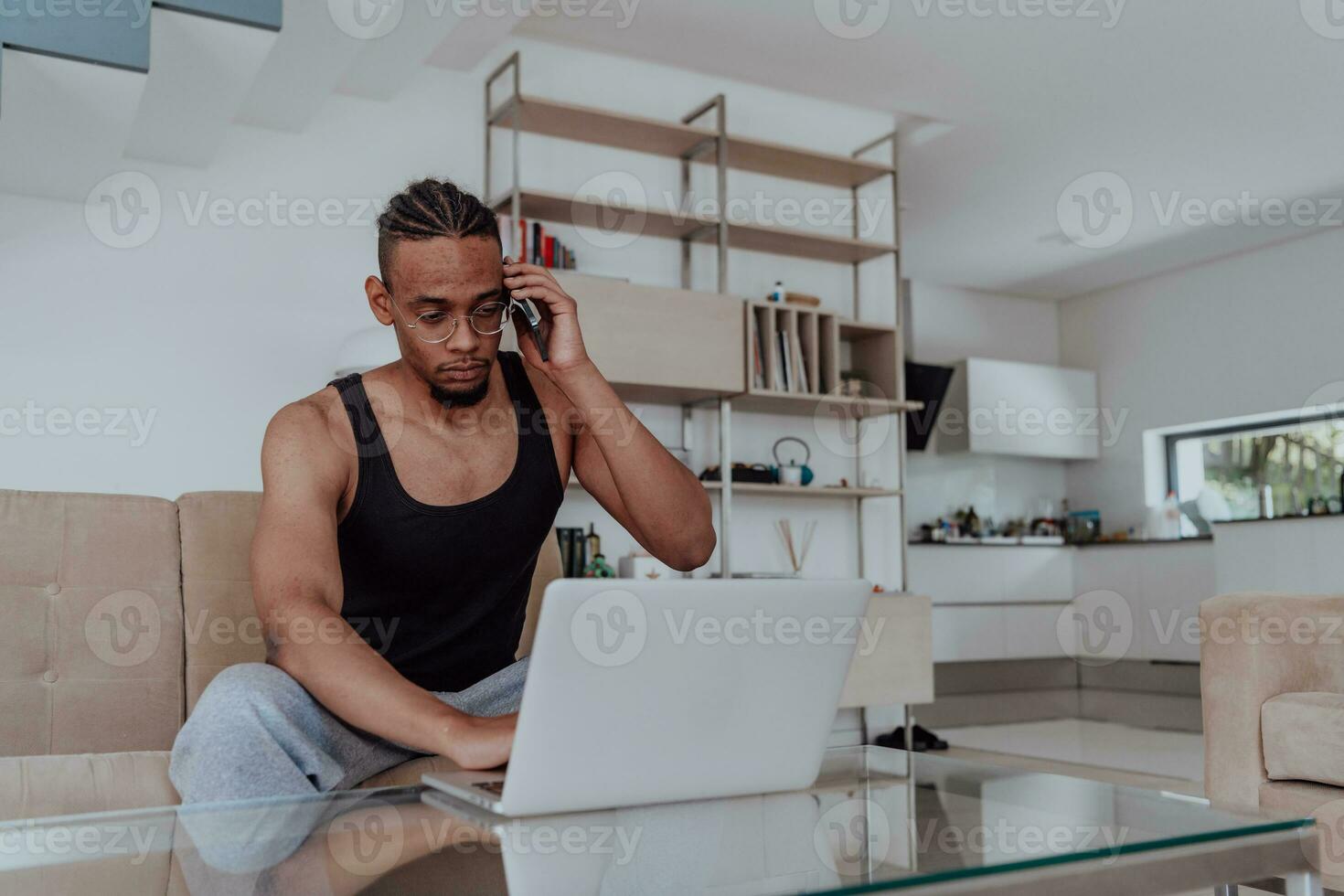 africano americano deporte hombre en lentes sentado a un mesa en un moderno vivo habitación, utilizando un ordenador portátil para negocio vídeo charlar, conversacion con amigos y entretenimiento foto