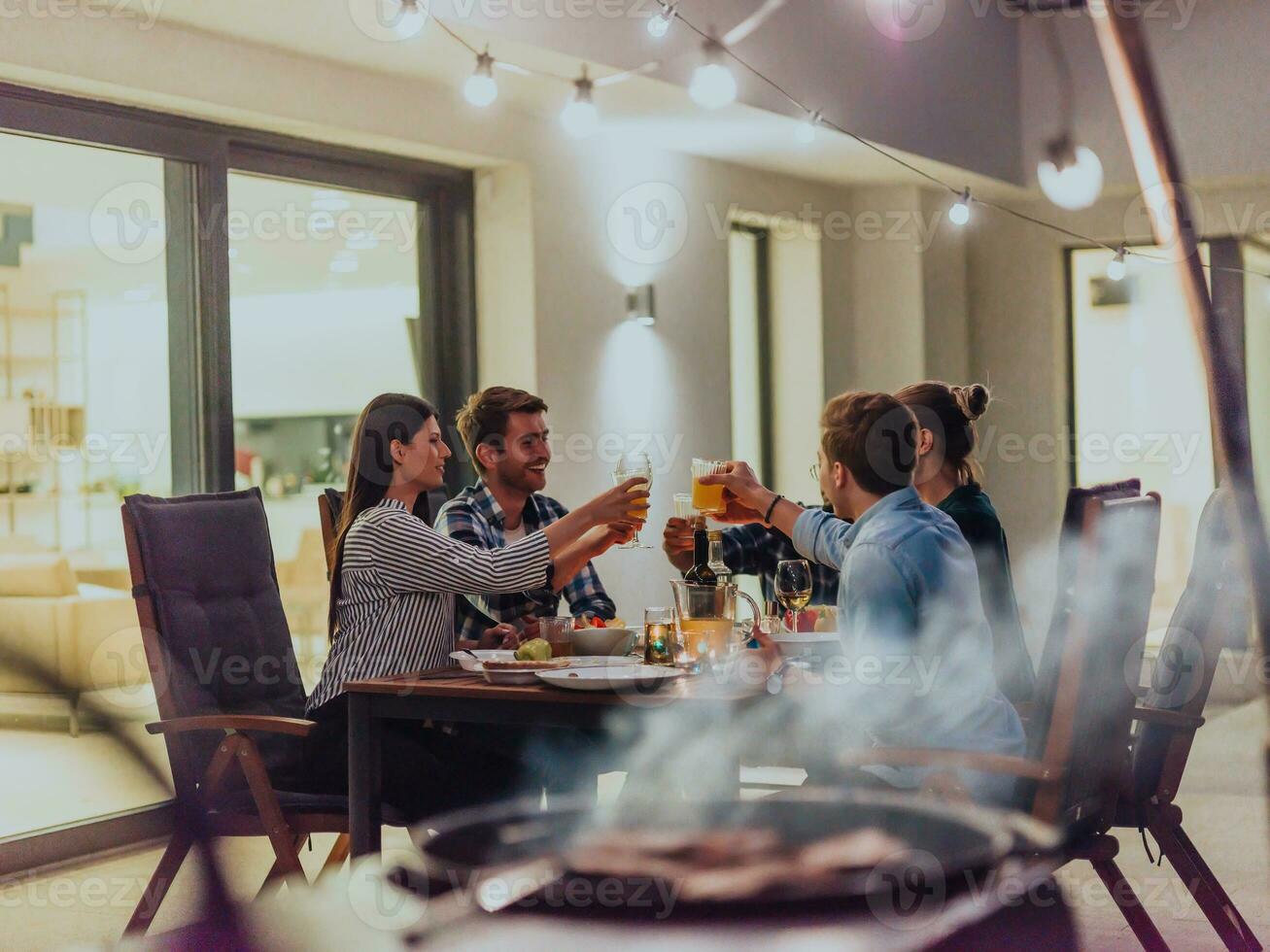 un grupo de joven diverso personas teniendo cena en el terraza de un moderno casa en el noche. divertido para amigos y familia. celebracion de vacaciones, bodas con parilla. foto