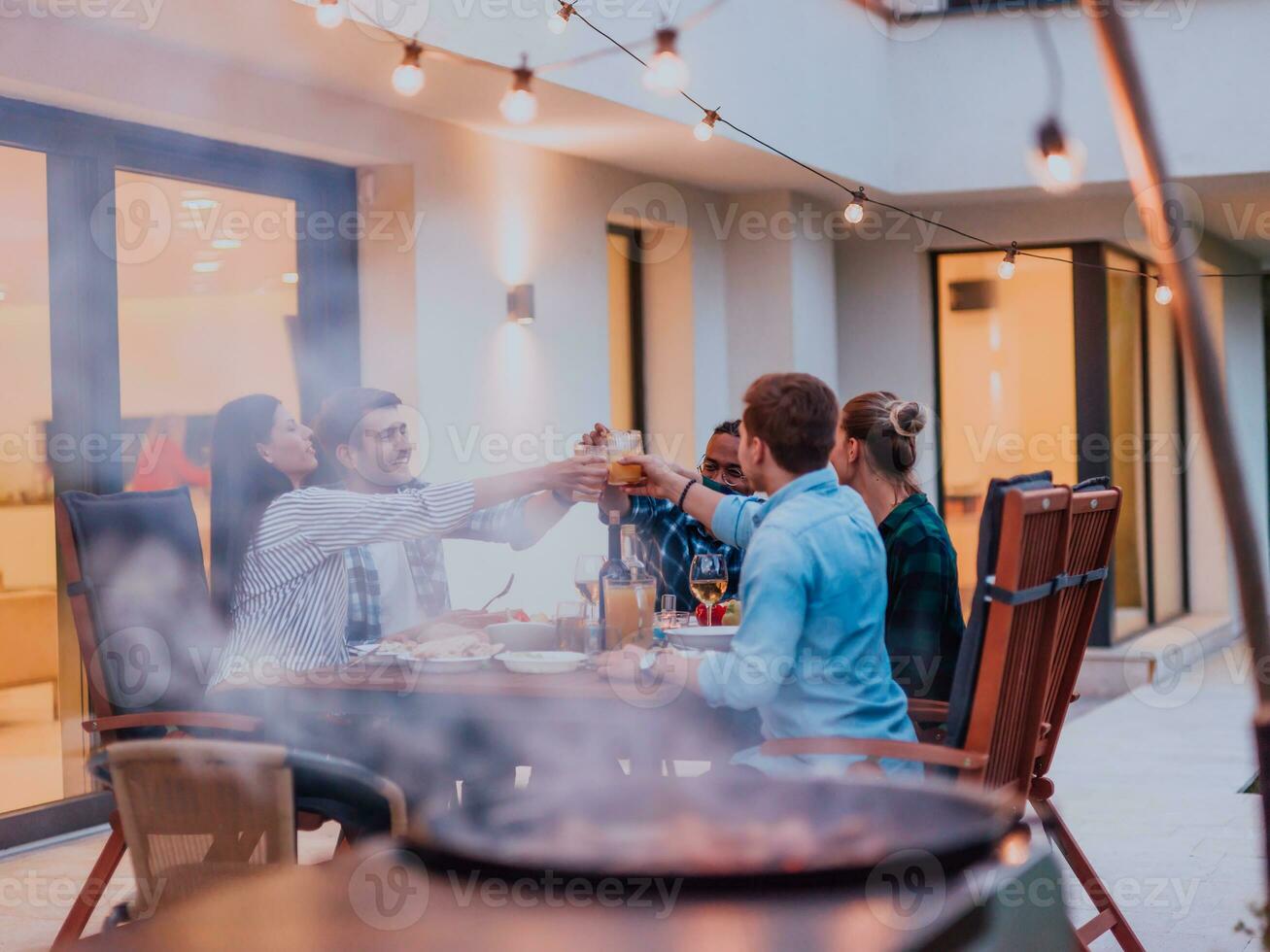 un grupo de joven diverso personas teniendo cena en el terraza de un moderno casa en el noche. divertido para amigos y familia. celebracion de vacaciones, bodas con parilla. foto