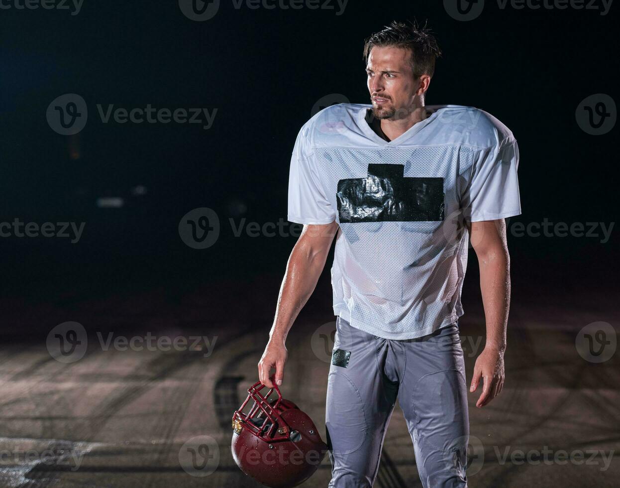 American Football Field Lonely Athlete Warrior Standing on a Field Holds his Helmet and Ready to Play. Player Preparing to Run, Attack and Score Touchdown. Rainy Night with Dramatic Fog, Blue Light photo