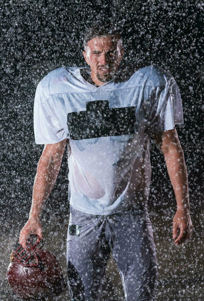 americano fútbol americano campo solitario atleta guerrero en pie en un campo sostiene su casco y Listo a jugar. jugador preparando a correr, ataque y Puntuación aterrizaje. lluvioso noche con dramático niebla, azul ligero foto