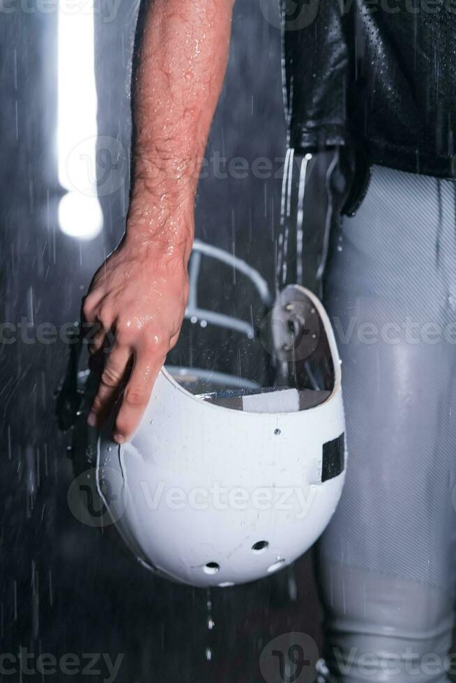 Close up of American Football Athlete Warrior Standing on a Field focus on his Helmet and Ready to Play. Player Preparing to Run, Attack and Score Touchdown. Rainy Night with Dramatic lens flare and rain drops. photo