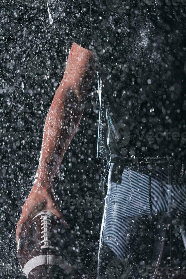 Close up of American Football Athlete Warrior Standing on a Field focus on ball and Ready to Play. Player Preparing to Run, Attack and Score Touchdown. Rainy Night with Dramatic lens flare and rain drops. photo