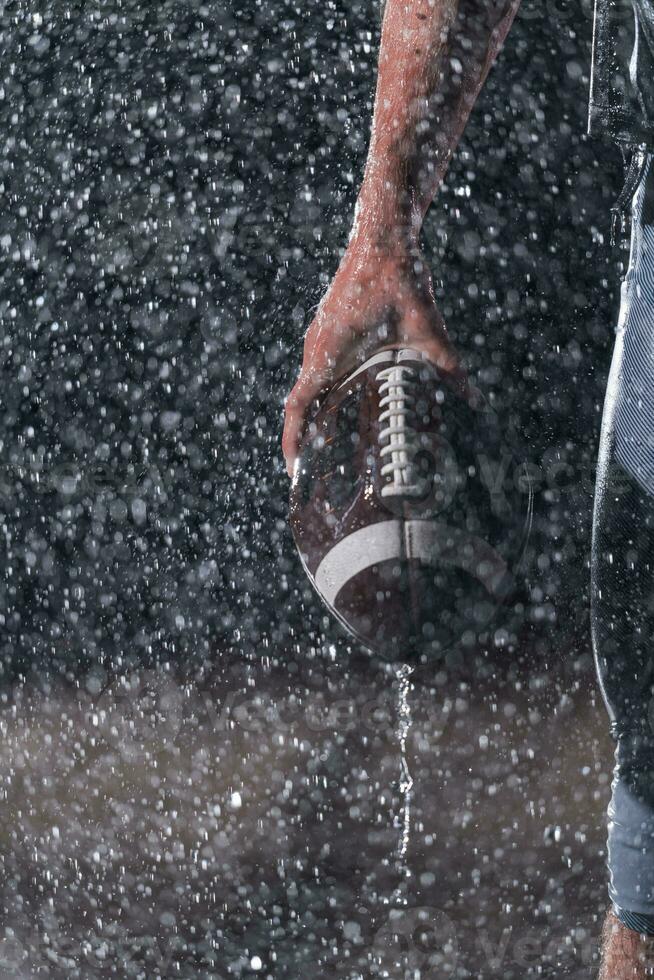 Close up of American Football Athlete Warrior Standing on a Field focus on ball and Ready to Play. Player Preparing to Run, Attack and Score Touchdown. Rainy Night with Dramatic lens flare and rain drops. photo