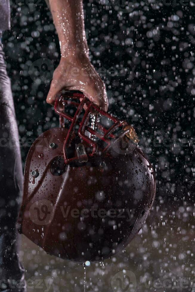 Close up of American Football Athlete Warrior Standing on a Field focus on his Helmet and Ready to Play. Player Preparing to Run, Attack and Score Touchdown. Rainy Night with Dramatic lens flare and rain drops. photo