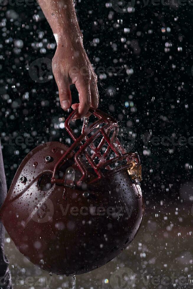 Close up of American Football Athlete Warrior Standing on a Field focus on his Helmet and Ready to Play. Player Preparing to Run, Attack and Score Touchdown. Rainy Night with Dramatic lens flare and rain drops. photo