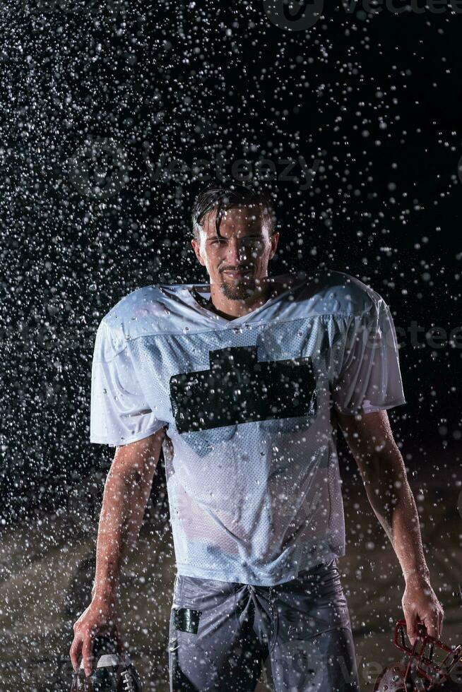 americano fútbol americano campo solitario atleta guerrero en pie en un campo sostiene su casco y Listo a jugar. jugador preparando a correr, ataque y Puntuación aterrizaje. lluvioso noche con dramático niebla, azul ligero foto