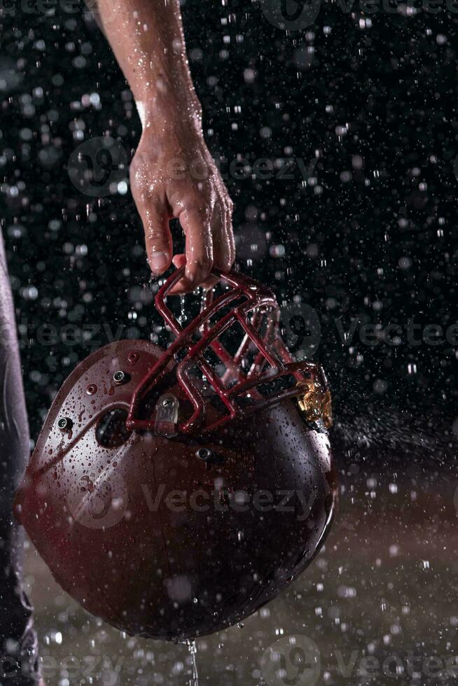 Close up of American Football Athlete Warrior Standing on a Field focus on his Helmet and Ready to Play. Player Preparing to Run, Attack and Score Touchdown. Rainy Night with Dramatic lens flare and rain drops. photo