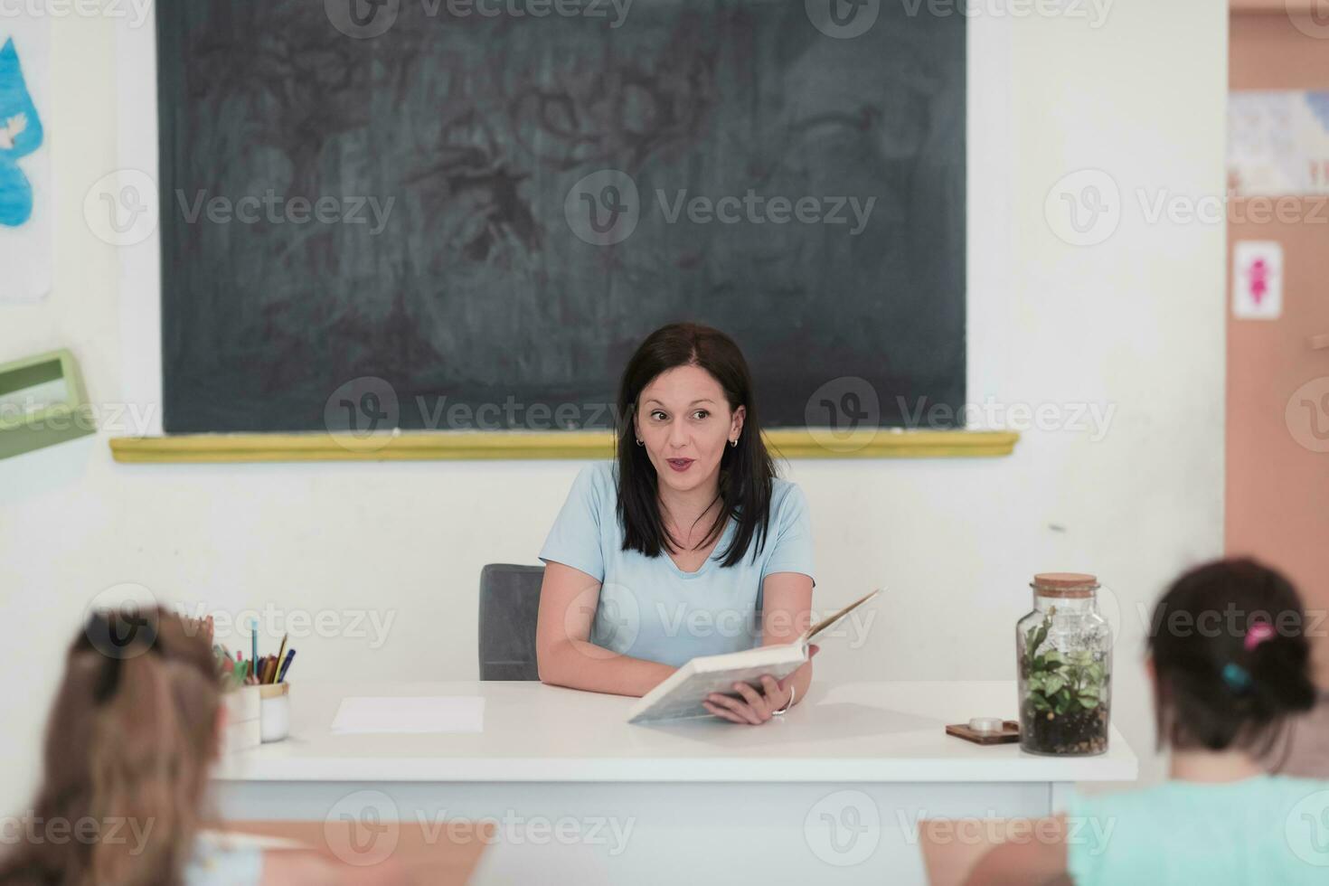 un profesor lee un libro a elemental colegio estudiantes quien escucha cuidadosamente mientras sentado en un moderno salón de clases foto