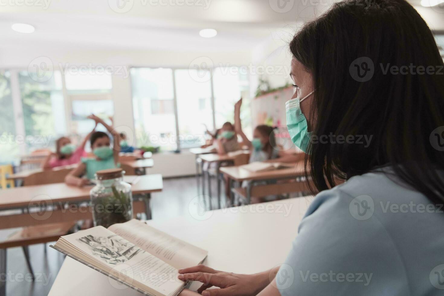 Multiracial group of kids wearing face masks working at class, writing and listening explanations of teacher in classroom photo