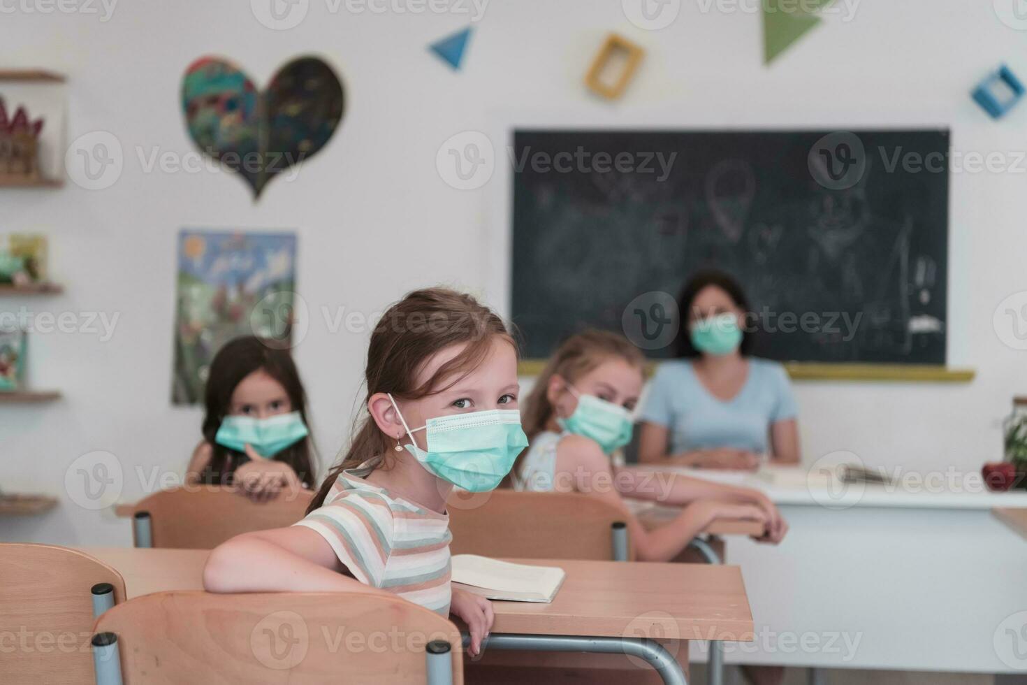 Multiracial group of kids wearing face masks working at class, writing and listening explanations of teacher in classroom photo