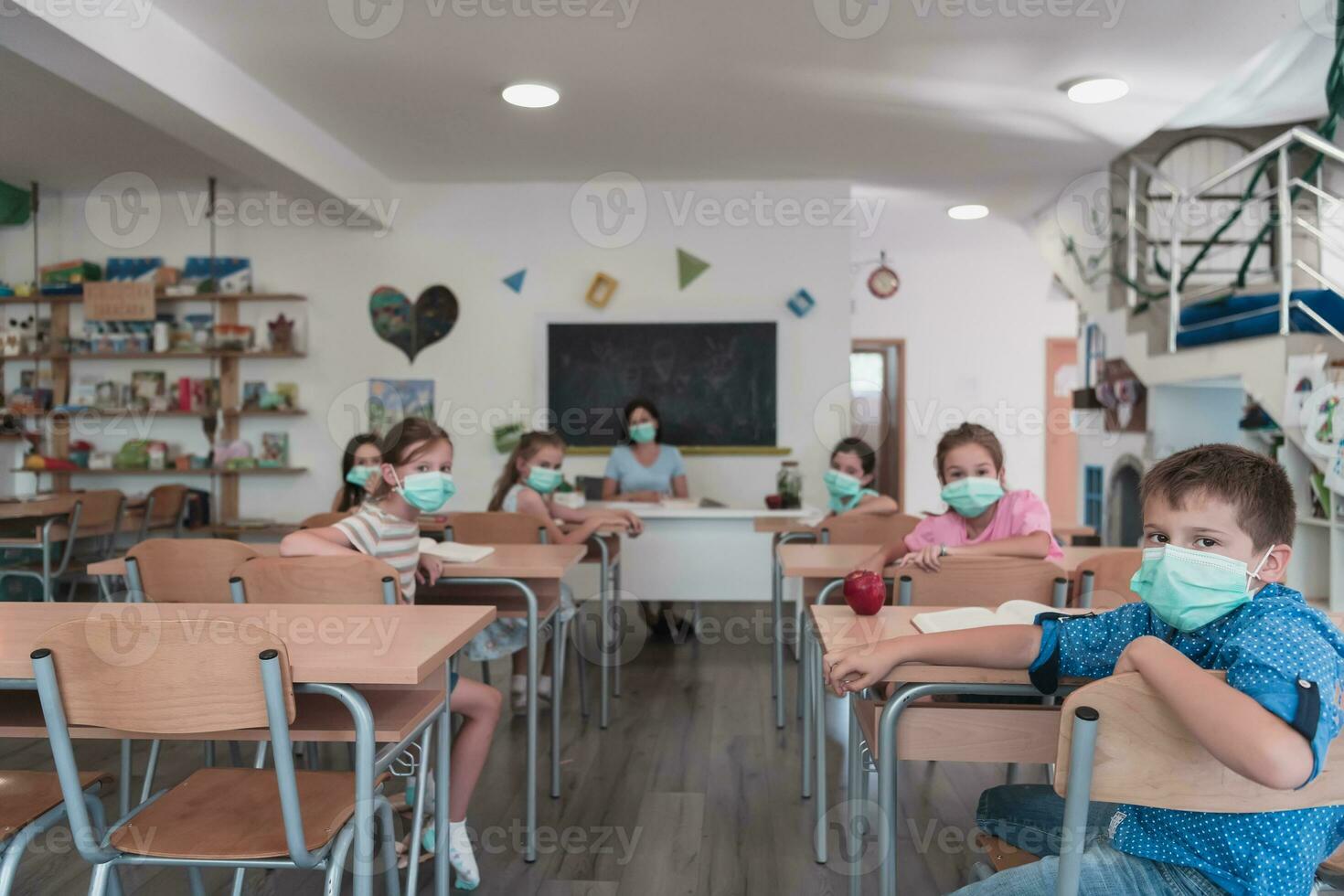 Multiracial group of kids wearing face masks working at class, writing and listening explanations of teacher in classroom photo