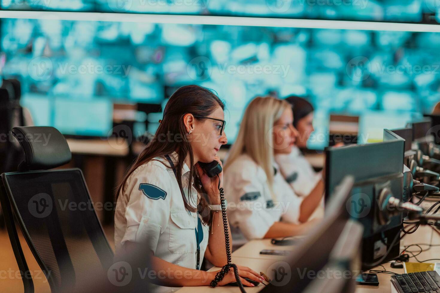 hembra seguridad Guardia operador hablando en el teléfono mientras trabajando a puesto de trabajo con múltiple muestra seguridad guardias trabajando en múltiple monitores foto