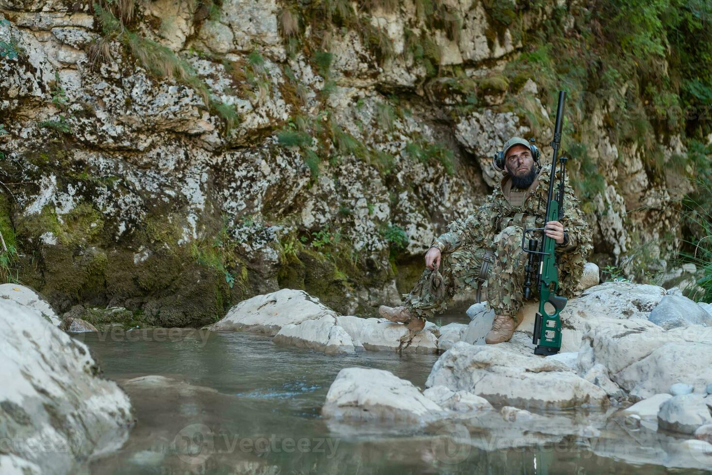 A military man or airsoft player in a camouflage suit sneaking the river and aims from a sniper rifle to the side or to target. photo