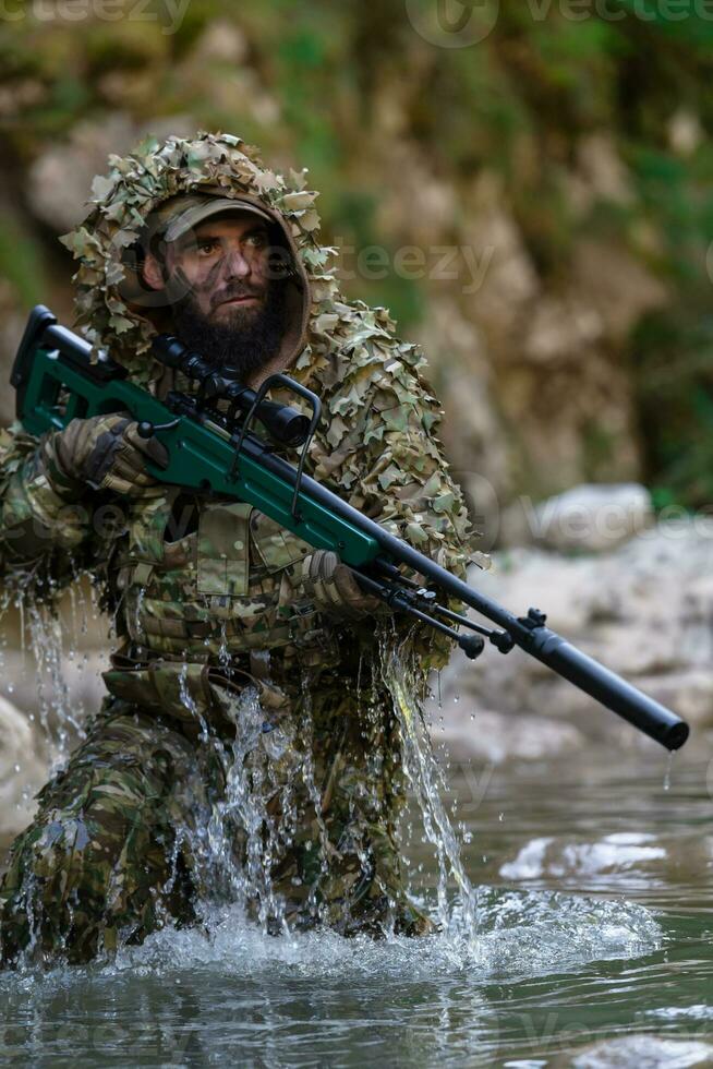 A military man or airsoft player in a camouflage suit sneaking the river and aims from a sniper rifle to the side or to target. photo