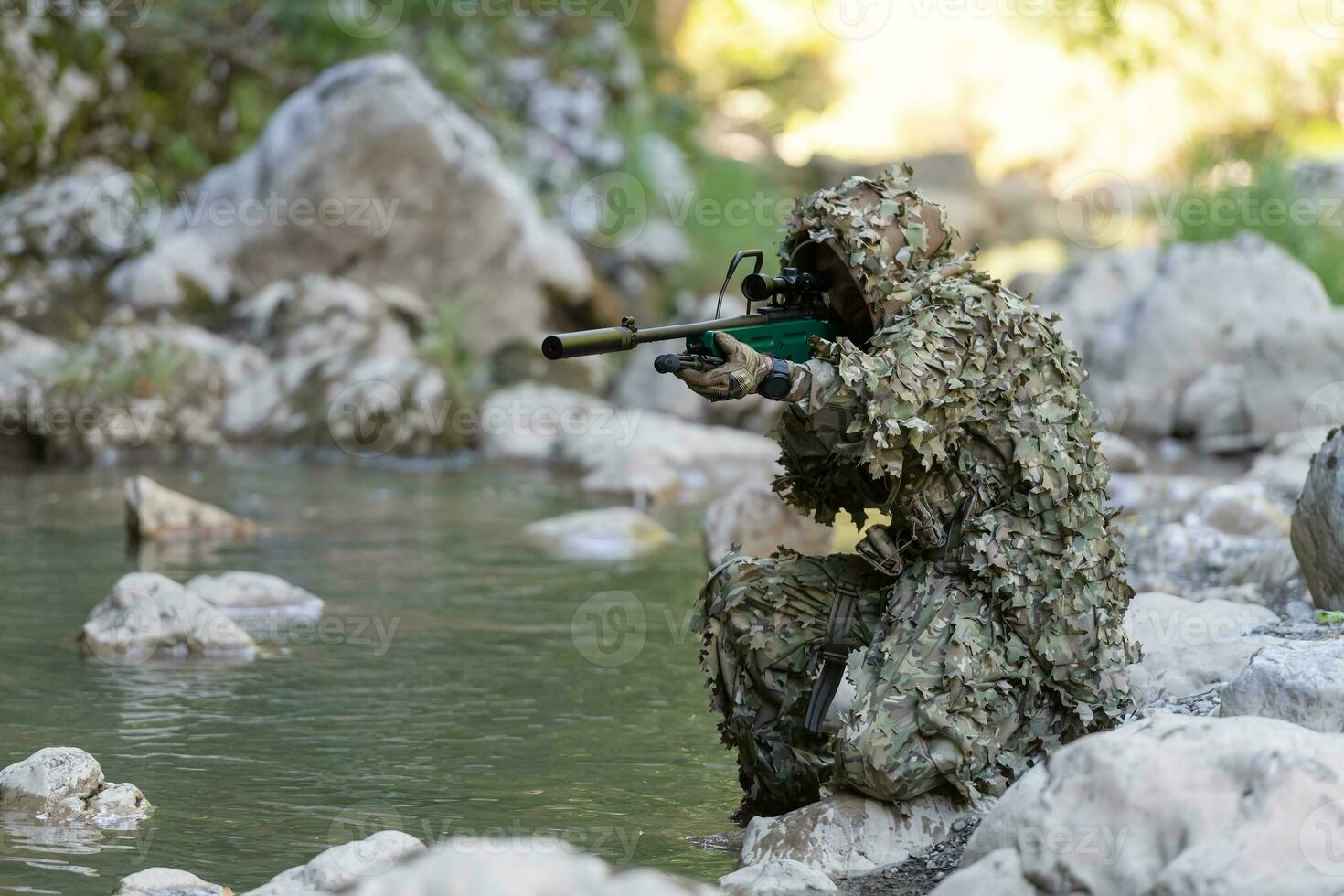 A military man or airsoft player in a camouflage suit sneaking the river and aims from a sniper rifle to the side or to target. photo