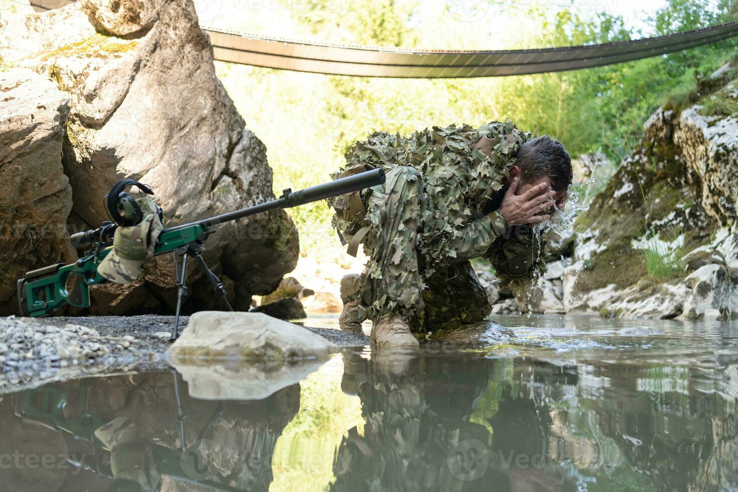 Soldier in a camouflage suit uniform drinking fresh water from the river. Military sniper rifle on the side. photo