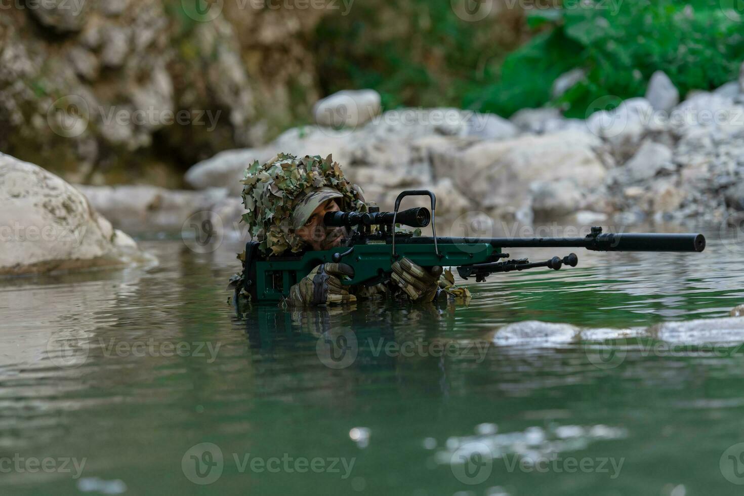 A military man or airsoft player in a camouflage suit sneaking the river and aims from a sniper rifle to the side or to target. photo