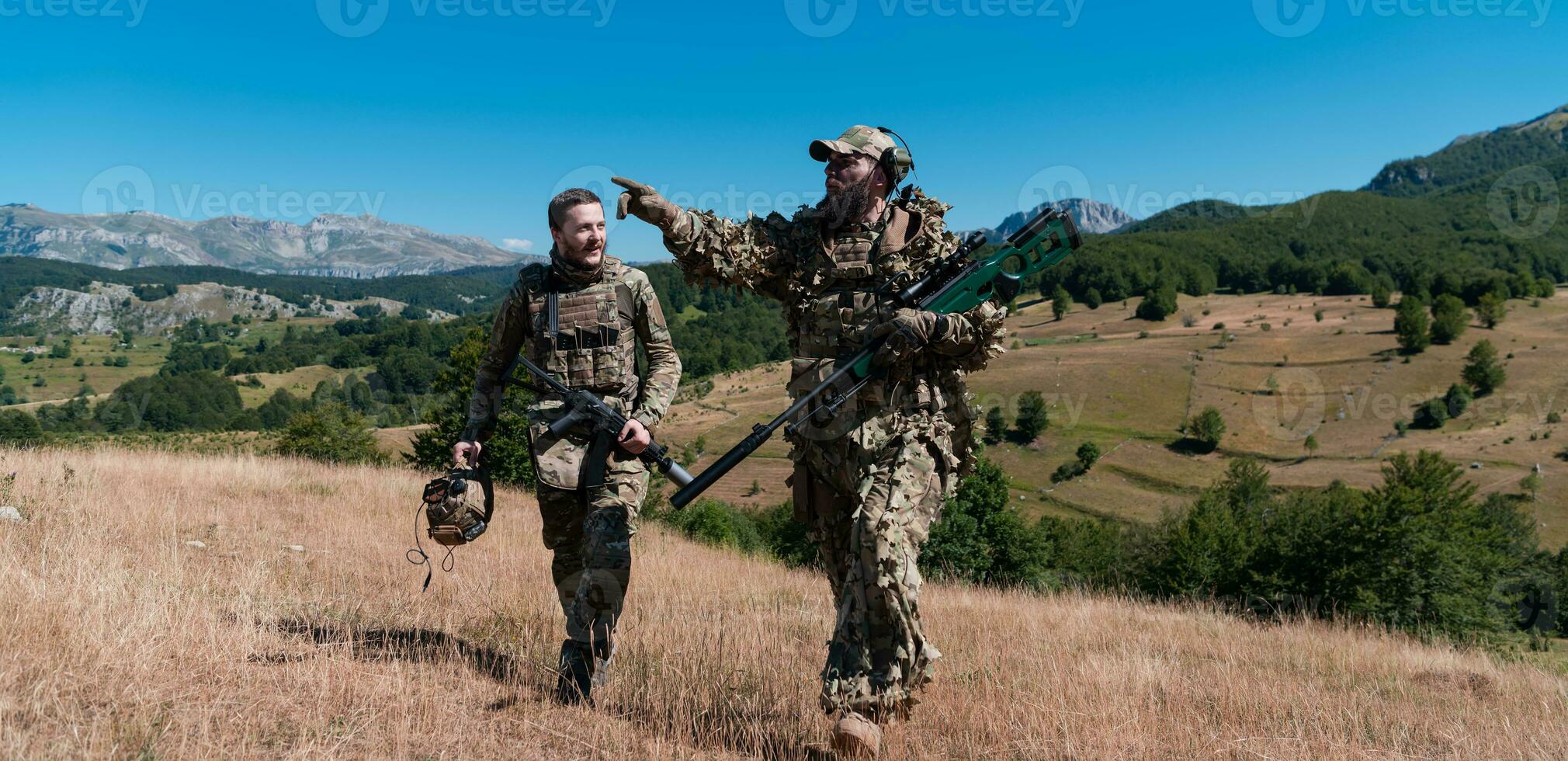 A sniper team squad of soldiers is going undercover. Sniper assistant and team leader walking and aiming in nature with yellow grass and blue sky. Tactical camouflage uniform. photo