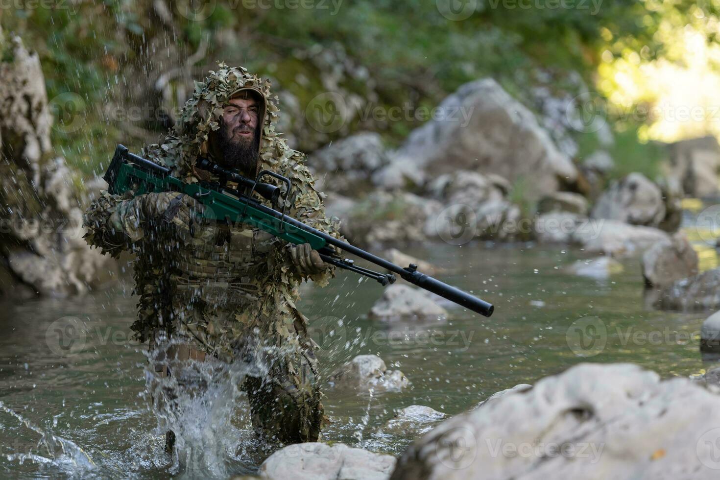 A military man or airsoft player in a camouflage suit sneaking the river and aims from a sniper rifle to the side or to target. photo