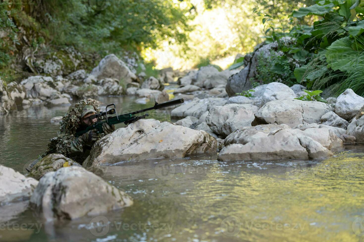 A military man or airsoft player in a camouflage suit sneaking the river and aims from a sniper rifle to the side or to target. photo