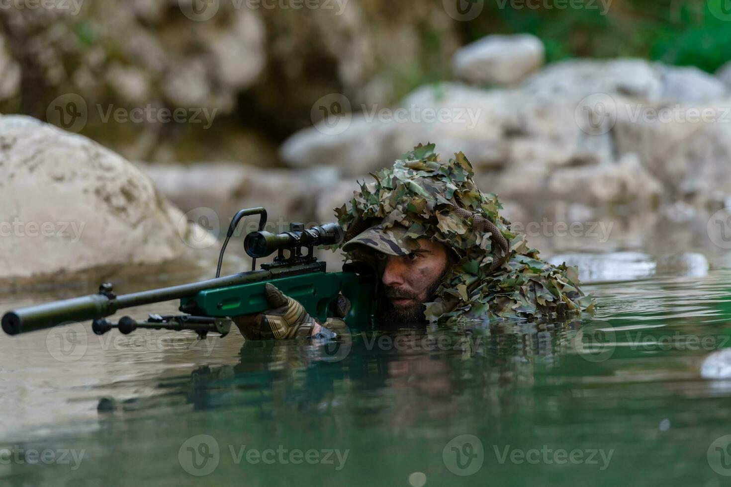 A military man or airsoft player in a camouflage suit sneaking the river and aims from a sniper rifle to the side or to target. photo