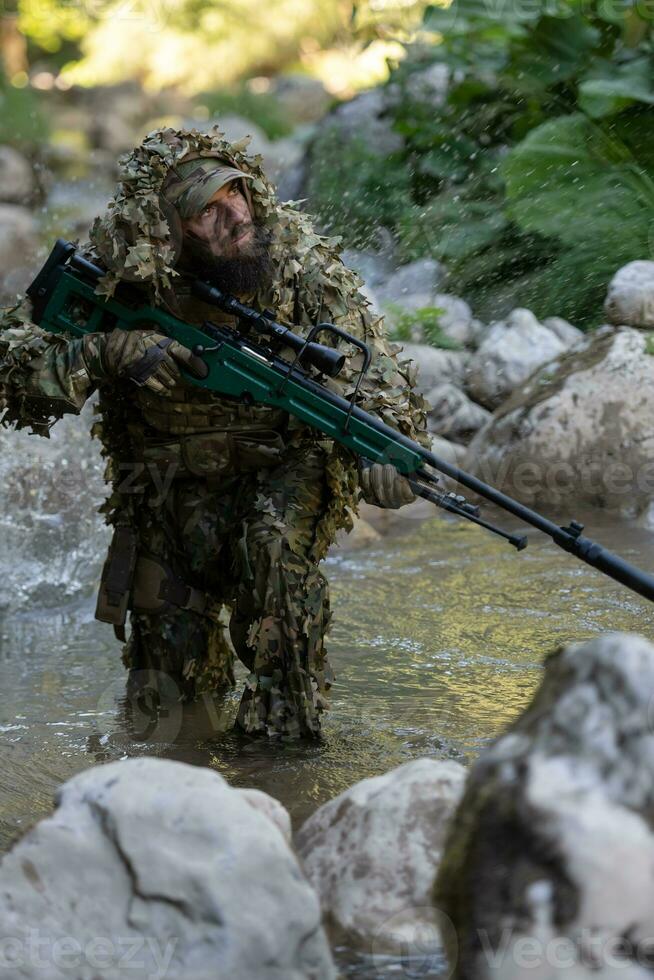 A military man or airsoft player in a camouflage suit sneaking the river and aims from a sniper rifle to the side or to target. photo