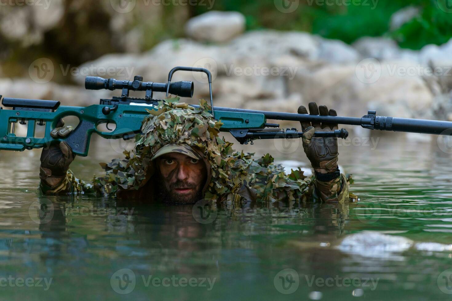 A military man or airsoft player in a camouflage suit sneaking the river and aims from a sniper rifle to the side or to target. photo