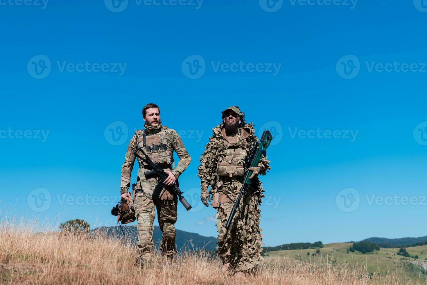 A sniper team squad of soldiers is going undercover. Sniper assistant and team leader walking and aiming in nature with yellow grass and blue sky. Tactical camouflage uniform. photo