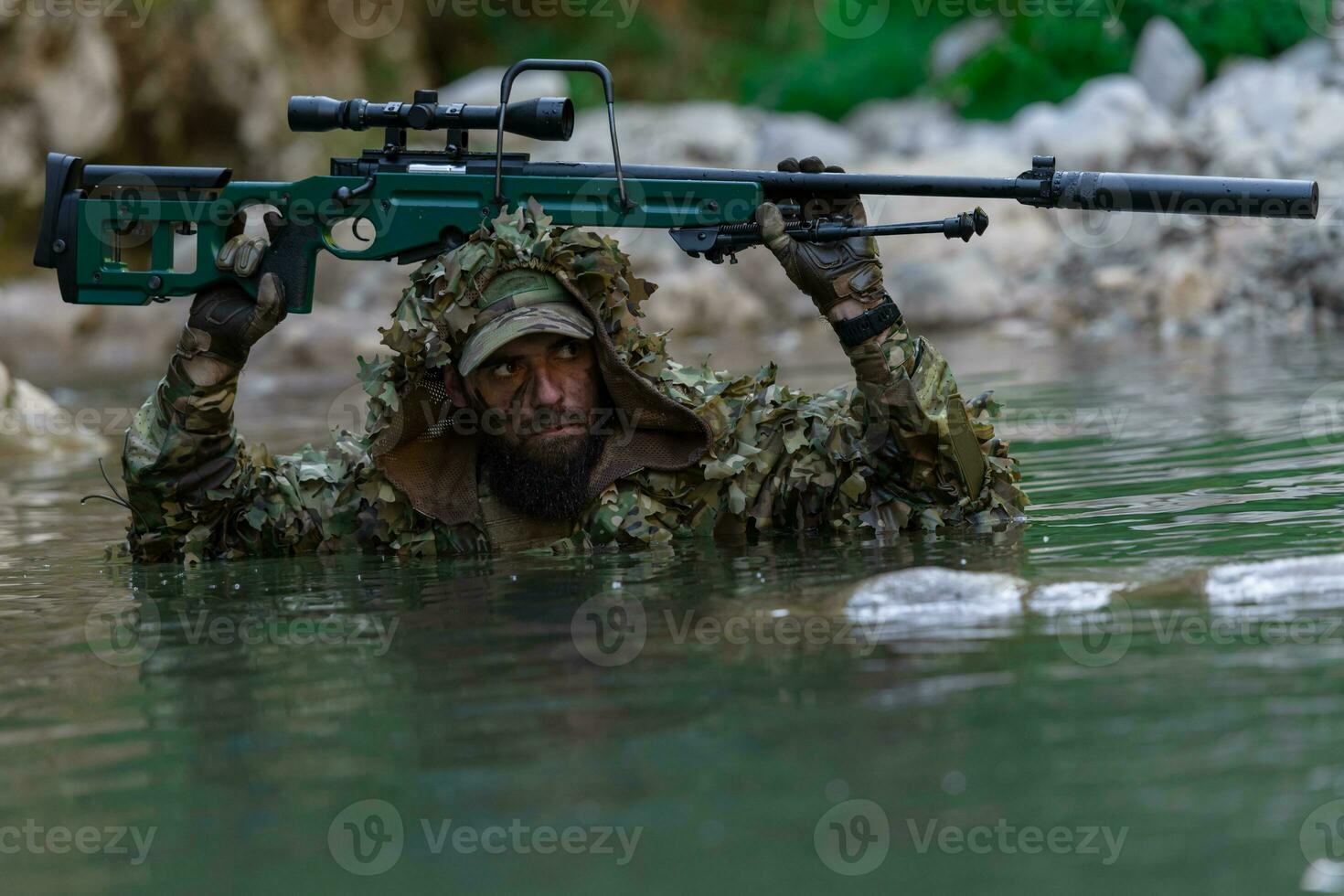 A military man or airsoft player in a camouflage suit sneaking the river and aims from a sniper rifle to the side or to target. photo