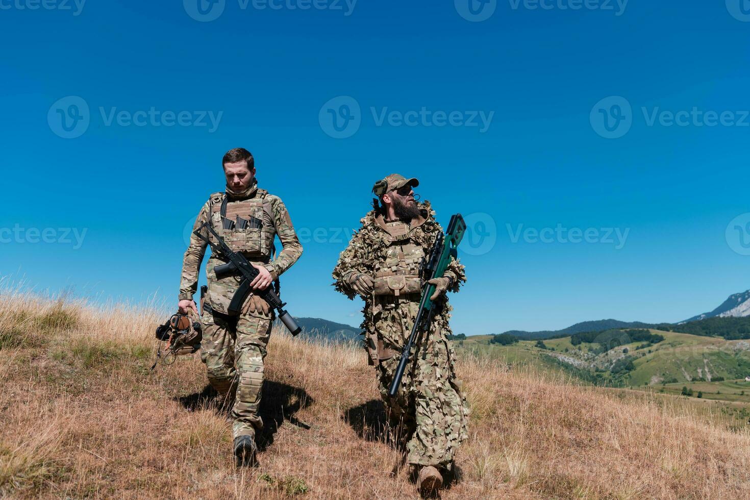 A sniper team squad of soldiers is going undercover. Sniper assistant and team leader walking and aiming in nature with yellow grass and blue sky. Tactical camouflage uniform. photo