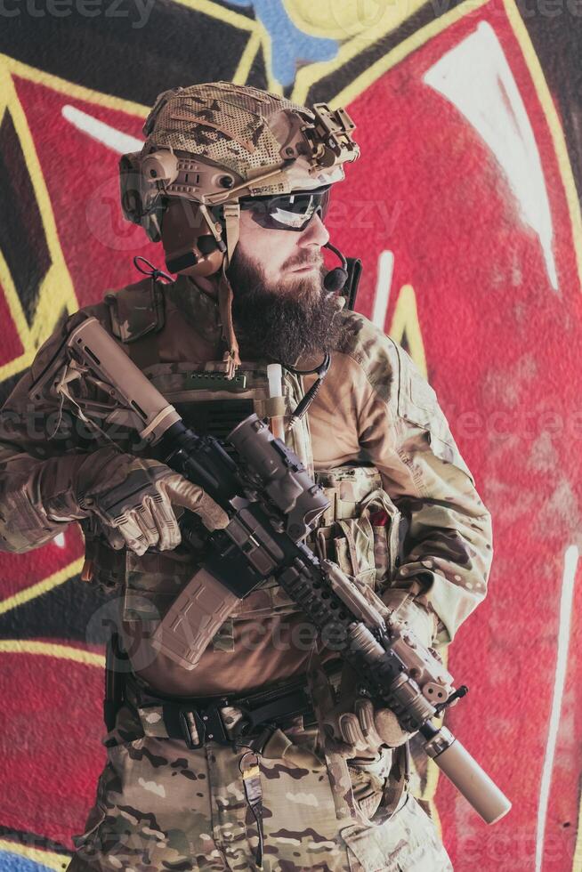 un barbado soldado en un especial efectivo uniforme soportes en frente de un dibujado pared con un rifle en su mano. selectivo atención foto