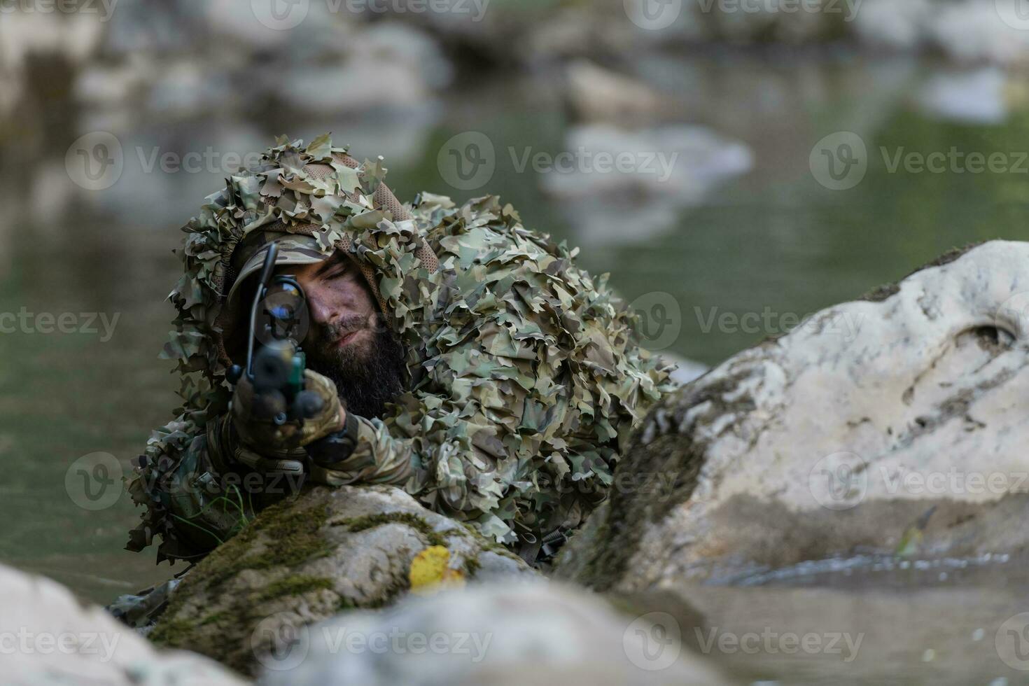 A military man or airsoft player in a camouflage suit sneaking the river and aims from a sniper rifle to the side or to target. photo