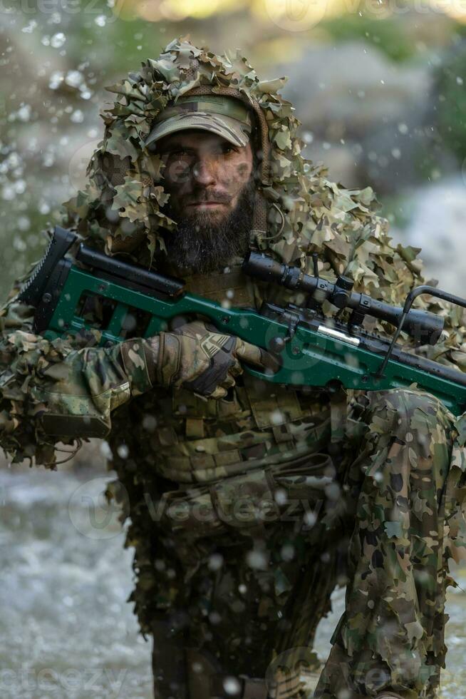 A military man or airsoft player in a camouflage suit sneaking the river and aims from a sniper rifle to the side or to target. photo