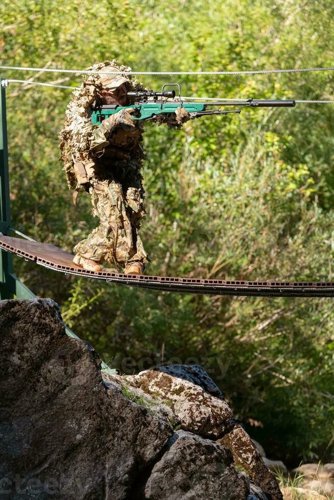 A military man or airsoft player in a camouflage suit sneaking the rope bridge and aims from a sniper rifle to the side or to target. photo
