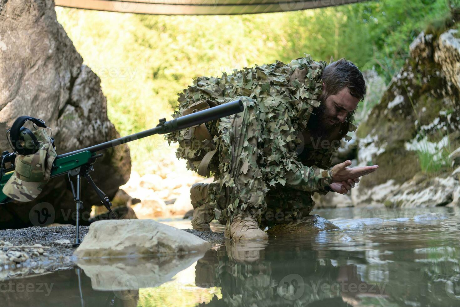 Soldier in a camouflage suit uniform drinking fresh water from the river. Military sniper rifle on the side. photo