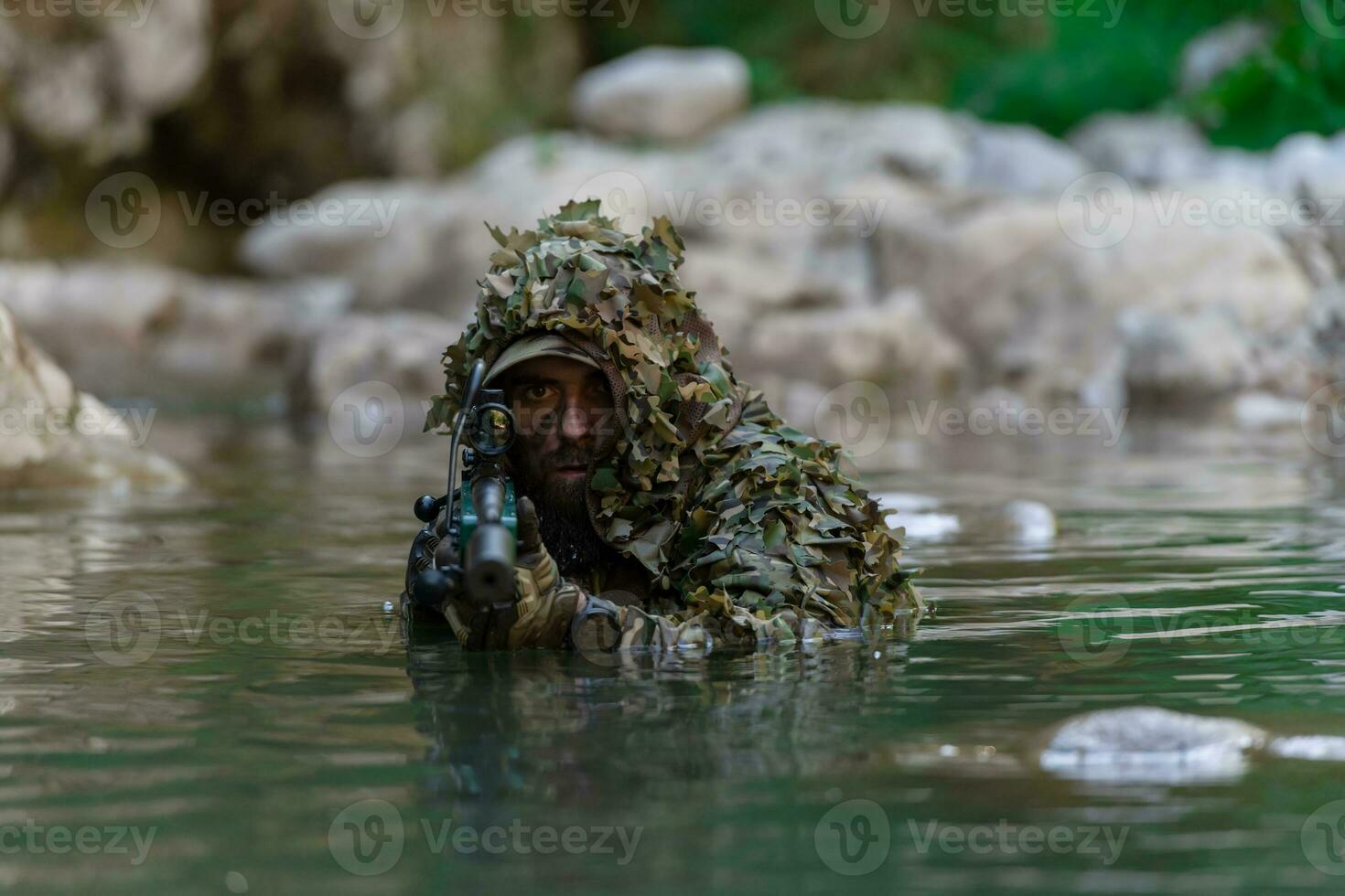 A military man or airsoft player in a camouflage suit sneaking the river and aims from a sniper rifle to the side or to target. photo