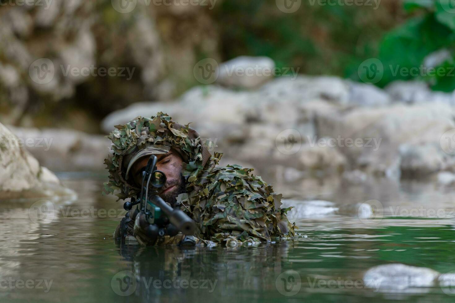 A military man or airsoft player in a camouflage suit sneaking the river and aims from a sniper rifle to the side or to target. photo