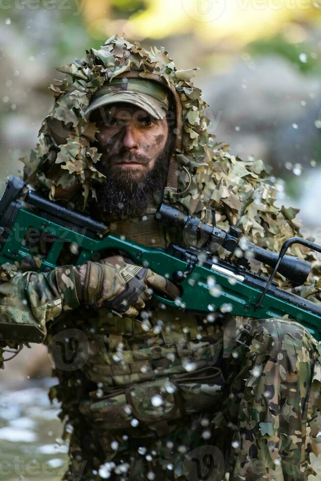A military man or airsoft player in a camouflage suit sneaking the river and aims from a sniper rifle to the side or to target. photo