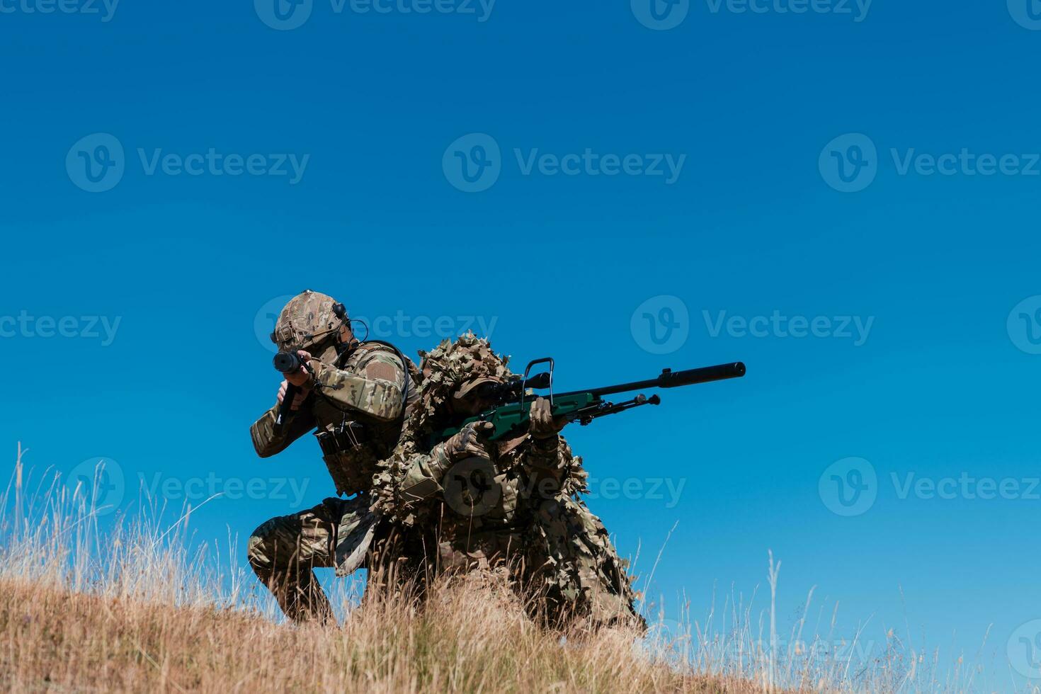 A sniper team squad of soldiers is going undercover. Sniper assistant and team leader walking and aiming in nature with yellow grass and blue sky. Tactical camouflage uniform. photo