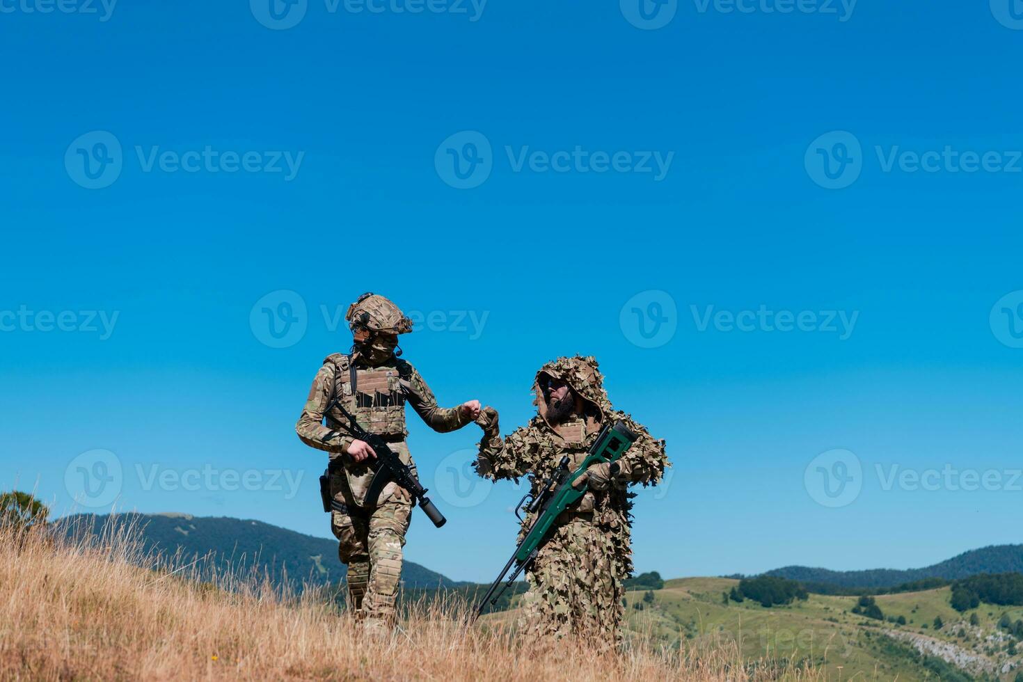 A sniper team squad of soldiers is going undercover. Sniper assistant and team leader walking and aiming in nature with yellow grass and blue sky. Tactical camouflage uniform. photo