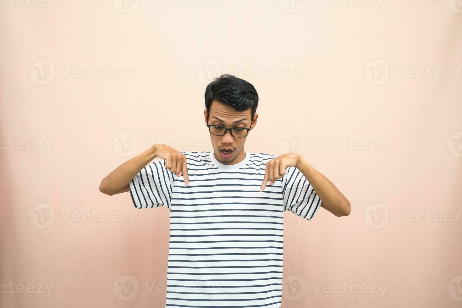 Asian man with glasses wearing casual striped shirt, whispering pose while pointing down. Isolated beige background. photo