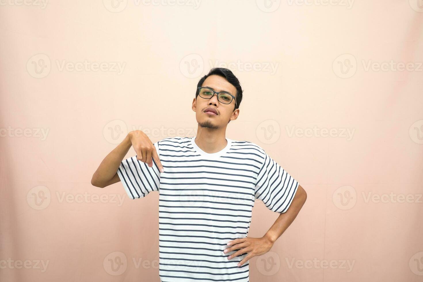 Asian man with glasses wearing casual striped shirt, whispering pose while pointing down. Isolated beige background. photo