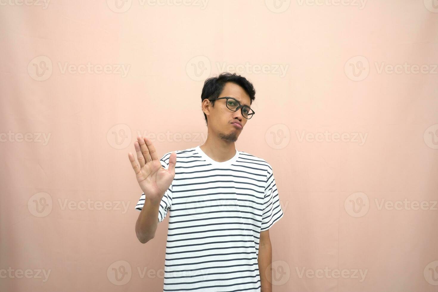 retrato de un asiático hombre en lentes vistiendo un casual a rayas camiseta. denotar detener, cruzar prohibición firmar, prohibir algo, rechazar,. beige antecedentes. foto