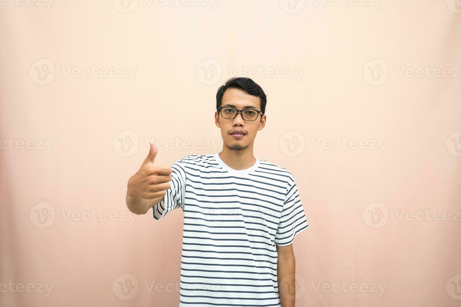 Asian man in glasses wearing casual striped shirt, posing giving thumbs up gesture or okay that agree. Isolated beige background. photo