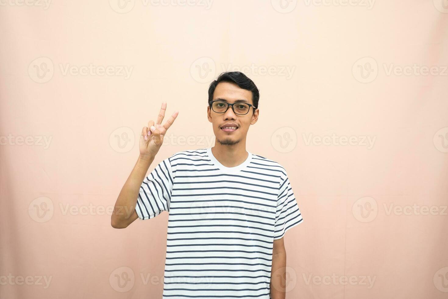 asiático hombre en lentes vistiendo casual a rayas camiseta, saludo actitud y sonriente. aislado beige antecedentes. foto