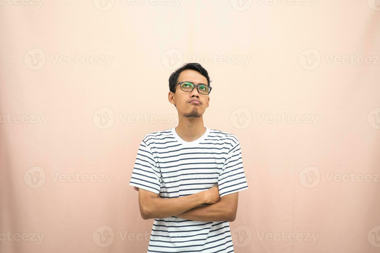 Asian man in glasses wearing casual striped t-shirt, gesturing get ideas, looking for ideas, thinking. Isolated beige background. photo