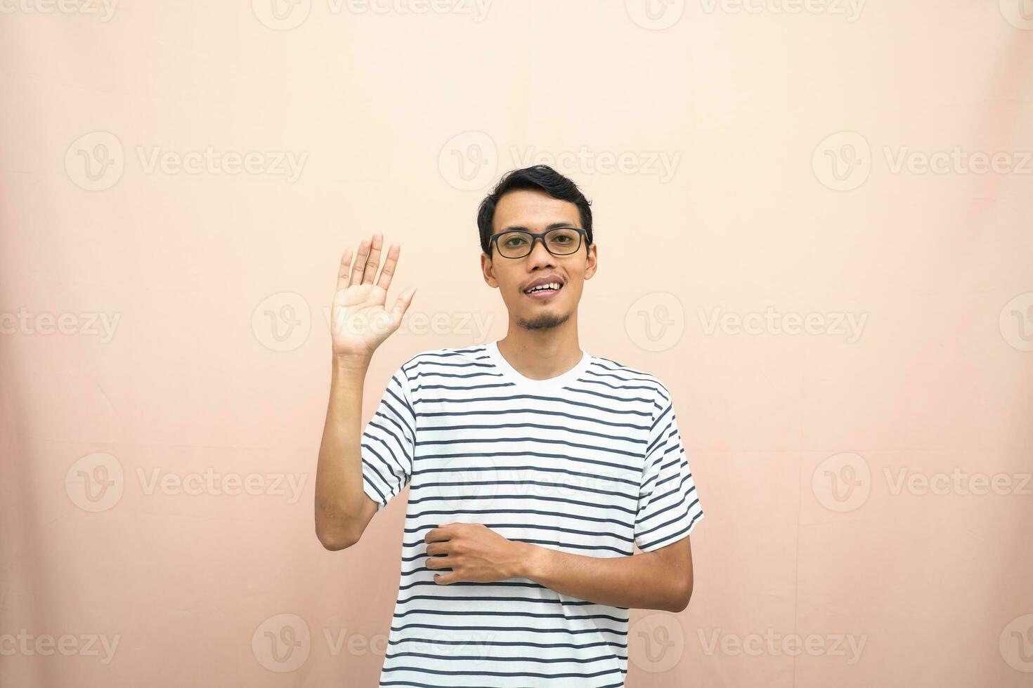 asiático hombre en lentes vistiendo casual a rayas camiseta, saludo actitud y sonriente. aislado beige antecedentes. foto